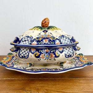 Puede incluir: Un plato de cerámica decorativo con tapa y bandeja a juego. El plato es de color crema con diseños florales azules, amarillos y naranjas. La tapa está rematada con un asa marrón en forma de piña. La bandeja tiene un borde festoneado.