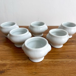 Può includere: Sei piccole ciotole in ceramica bianca con corpi arrotondati e piccole maniglie. Le ciotole sono disposte su una superficie di legno. Le ciotole hanno un design semplice ed elegante, adatto per servire piccole porzioni.