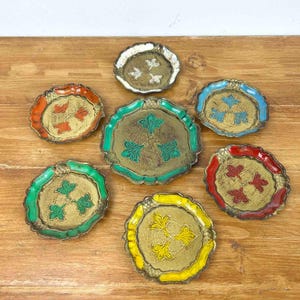 May include: A collection of decorative, vintage-style trays with scalloped edges. Each tray features a gold-colored center with a floral design and a colored border in shades of red, orange, green, blue, and yellow. The trays are arranged on a wooden surface.