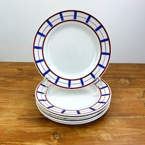 May include: A stack of white ceramic plates with a red and blue geometric border design. The plates are stacked on a wooden surface. The plates have a clean, minimalist aesthetic.