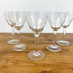 Op de afbeelding: Een set van vijf heldere wijnglazen met geëtste details. De glazen hebben lange stelen en afgeronde kelken. Ze staan op een houten oppervlak, wat hun elegante ontwerp benadrukt. Elk glas is ongeveer 15 cm hoog.
