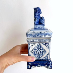 Può includere: Un contenitore in ceramica blu e bianco con una statuina decorativa a forma di leone sulla parte superiore. Il contenitore ha una forma quadrata con motivi ornati e una base a piedistallo. Il design presenta una combinazione di colori blu e bianco, creando un'estetica classica.