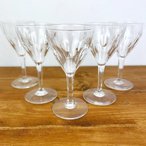 May include: A set of five clear crystal wine glasses with a decorative etched pattern. The glasses have a slender stem and a round base, set on a wooden surface.