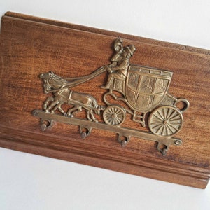 May include: A vintage key holder with a brass carriage and horse design mounted on a wooden board. The brass carriage features intricate details, and the board has four hooks for keys.