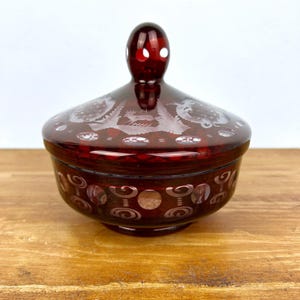 Könnte beinhalten: Ein rubinrotes Glasgefäß mit Deckel. Das Gefäß weist geätzte Blumen- und Scrollmuster auf. Der Deckel hat einen abgerundeten Griff mit weißen Akzenten. Das Gefäß steht auf einer Holzoberfläche.