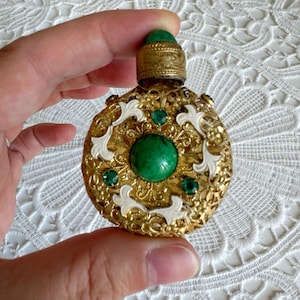Op de afbeelding: Een rond parfumflesje van goudkleurig metaal met ingewikkeld filigraanwerk, witte emaille accenten en een grote centrale groene cabochonsteen. Het flesje is versierd met kleinere groene gefacetteerde steentjes en een bijpassende groene dop.