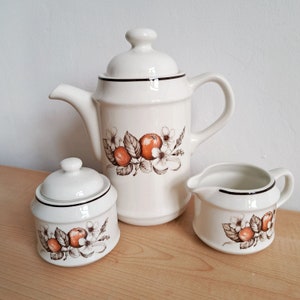 Peut inclure: Service à thé en céramique blanche comprenant une théière avec couvercle, un sucrier avec couvercle et un pot à crème. Chaque pièce est ornée d'un motif floral et fruité aux tons orange, marron et blanc, avec des bordures noires.