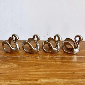 May include: Four silver swan-shaped napkin rings. Each ring features a stylized swan design with a curved neck and open ring base. The napkin rings are arranged in a row on a wooden surface.