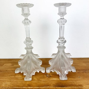 Könnte beinhalten: Zwei Kerzenhalter aus mattiertem Glas. Jeder hat ein detailliertes, verziertes Design mit breitem, blattartigem Fuß, schlankem Stiel und gewellter Oberseite. Die Kerzenhalter stehen auf einer Holzoberfläche.