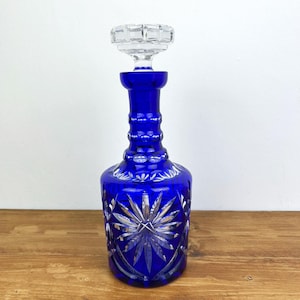 Puede incluir: Una jarra de vidrio azul cobalto con un tapón de vidrio transparente. La jarra presenta un diseño de estrella en la base y un cuello texturizado. El tapón tiene forma geométrica. La jarra está sobre una superficie de madera.