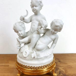 Puede incluir: Figurita de porcelana blanca con tres figuras de querubines. Uno de los querubines sostiene un objeto en forma de media luna. La figura descansa sobre una base dorada con detalles ornamentales, añadiendo un toque de elegancia. La base mide unos 10 cm de diámetro.