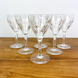 May include: Six clear crystal wine glasses with a textured, diamond-like pattern on the bowl. The glasses have long stems and round bases, arranged on a wooden surface. The glasses are empty and reflect the light.