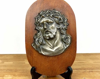 Könnte beinhalten: Religiöse Wandplakette mit einem Metallrelief eines Mannes mit Bart und Dornenkrone, montiert auf einem ovalen Holz. Das Holz hat einen warmen, rötlich-braunen Farbton. Die Plakette wird auf einem schwarzen Ständer ausgestellt.