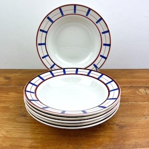 May include: A set of white ceramic bowls with a decorative design. Each bowl features a red rim with blue vertical stripes and a red inner border. The bowls are stacked on a wooden surface.