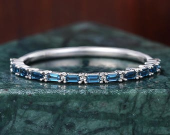 May include: A silver band ring with alternating blue rectangular gemstones and small clear stones. The ring is set on a dark green marble surface, with a blurred brown background.