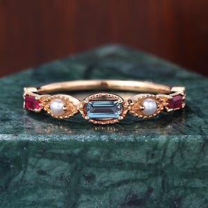 Może przedstawiać: Złoty pierścionek z centralnym kamieniem akwamarynowym w kształcie bagietki, otoczonym czerwonymi kamieniami szlachetnymi i małymi perłami. Pierścionek ma bogaty wzór z delikatną obrączką. Kamienie szlachetne osadzone są w złotej oprawie.