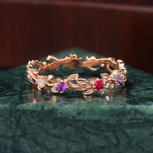 May include: A rose gold ring with a floral design. The ring features an opal, amethyst, and ruby gemstone. The ring is set on a green surface. The ring is a delicate piece of jewelry.