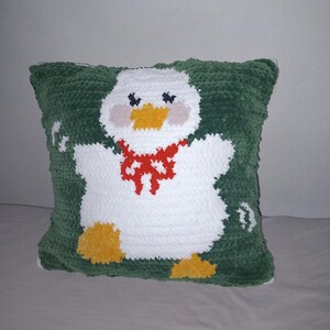 May include: A handmade, square pillow with a crocheted duck design. The duck is white with a yellow beak and feet, pink cheeks, and a red bow. The pillow's background is green, and the pillow measures approximately 14 inches square.