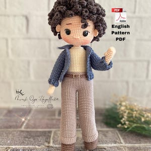Könnte beinhalten: Handgefertigte Häkelpuppe mit braunen Locken, die eine blaue Jacke, ein beiges Hemd und eine braune Hose trägt. Die Puppe hat einen Gürtel und steht auf einer Ziegeloberfläche. Der Text sagt "Adobe English Pattern PDF".