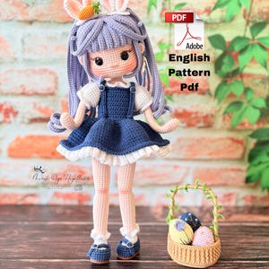 May include: A handmade crochet doll with a blue dress, white ruffled trim, and a bunny ear headband with a carrot. The doll has purple hair and is holding a small basket with Easter eggs. The image includes the text "Adobe English Pattern Pdf".
