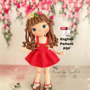 Könnte beinhalten: Häkelpuppe mit braunen Haaren, einem roten Kleid und einer Blumenkrone. Die Puppe steht auf einer weißen Oberfläche mit einem rosa floralen Hintergrund. Der Text "English Pattern PDF" ist im Bild sichtbar.