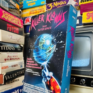 May include: A VHS tape of the horror movie "Killer Klowns from Outer Space". The cover features a clown with a red and white costume, holding a weapon that looks like a giant ice cream cone. The text "Killer Klowns from Outer Space" is in pink and black letters.
