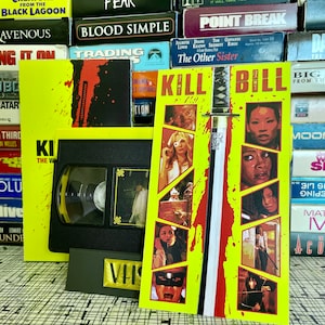 May include: A yellow and red VHS tape cover with the title "KILL BILL" in black text. The cover features a sword and multiple images of a woman in different scenes from the movie.
