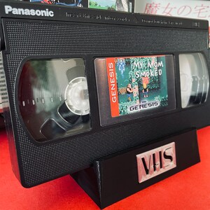 May include: A black VHS tape with a label that reads "My Mom Smoked" and a Sega Genesis logo. The tape is sitting on a black stand with the word "VHS" printed on it.