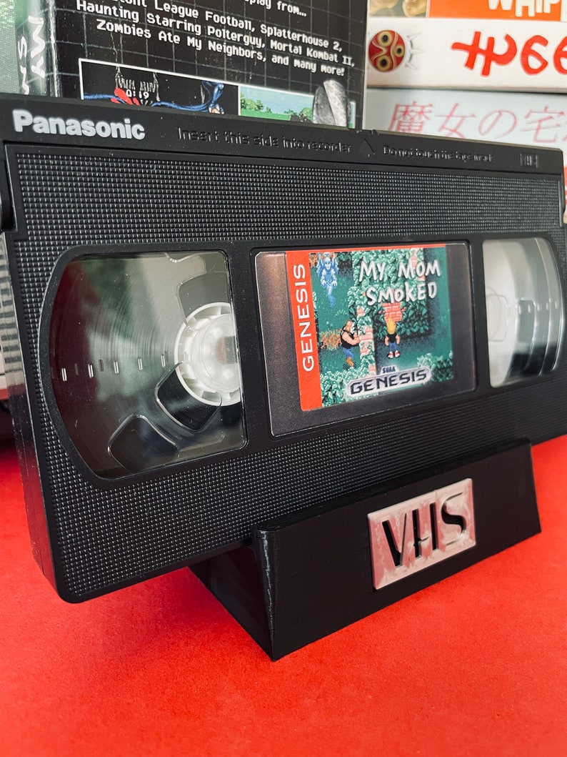 May include: A black VHS tape with a label that reads "My Mom Smoked" and a Sega Genesis logo. The tape is sitting on a black stand with the word "VHS" printed on it.