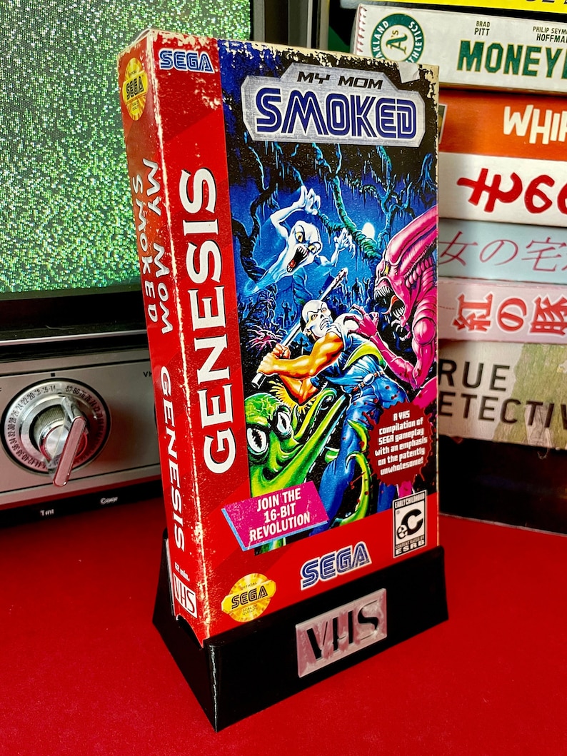 May include: A Sega Genesis game cartridge for the game "My Mom Smoked" with a black and white illustration of a character holding a weapon and a green monster on the front. The cartridge is in a black plastic VHS case with the word "VHS" printed on the front.