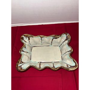 May include: Rectangular ceramic serving dish with a wavy rim. The dish has a light beige interior and is trimmed with brown and turquoise accents. The rim has a brown glaze with turquoise streaks. The dish is on a red surface.