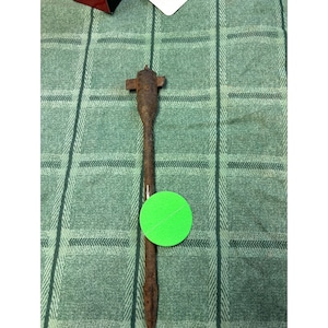 May include: A rusty metal tool with a pointed end and a cross-shaped head. The tool is lying on a green and white checkered surface.