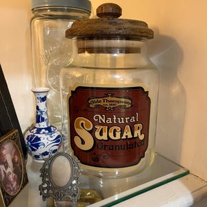 May include: A clear glass storage jar with a dark wooden lid. The jar features a rectangular label with a dark red background and cream-colored text that reads Olde Thompson est. 1887 Natural Sugar Granulated, with the word Sugar in large font.