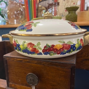 May include: A white enameled metal casserole dish with a matching lid, featuring a colorful fruit pattern of red apples, green pears, and dark blue berries. The pot has two gold-toned metal handles and a gold-toned knob on the lid's center.
