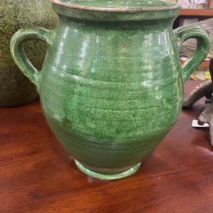 May include: A green ceramic vessel featuring a wide mouth, a rounded body, and two side handles. The exterior surface displays a distinct crackle glaze finish with variations in color intensity, and the rim shows a thin, brownish edge.