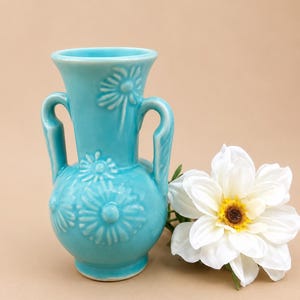 May include: A turquoise ceramic vase with two side handles and a raised floral pattern featuring daisy-like flowers. The vase has a flared rim and a rounded body, accompanied by a single white artificial flower with a yellow and brown center.