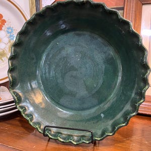 May include: A round, deep-dish ceramic pie plate with a dark forest green glaze and a glossy finish. The rim features a decorative, scalloped, or fluted edge design. The interior surface is smooth, showing subtle variations in the green color tone.