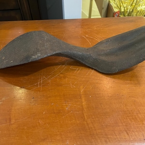 May include: A dark gray, weathered cast iron shoe cobbler's anvil or last. The object features a smooth, elongated, and curved shape with a wide, rounded heel section that tapers into a narrower, slightly pointed toe area, showing a textured surface.