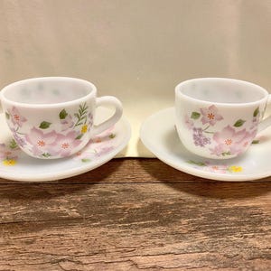 May include: Two white teacups and saucers with floral designs. The cups and saucers are decorated with pink, purple, and yellow flowers and green leaves. The cups have a handle and sit on matching saucers.
