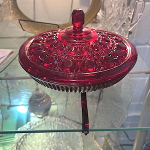 May include: A round, ruby red glass candy dish with a matching lid. The lid features a faceted, geometric hobnail pattern and a central knob handle. The base of the dish has vertical ribbing along the exterior edge, creating a textured appearance.
