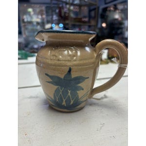 May include: A small, brown ceramic pitcher with a blue pineapple design. The pitcher has a handle and a spout.