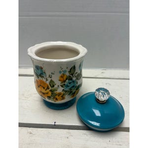 May include: A white ceramic jar with a floral design in yellow and blue, and a teal base. The jar has a matching teal lid with a clear, crystal-like knob. The jar is likely for storage or decoration.
