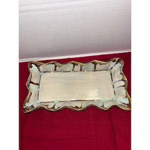 May include: Rectangular ceramic serving tray with a wavy, brown-edged border. The tray has a cream-colored interior and light blue drips along the edge. The tray is on a red surface.
