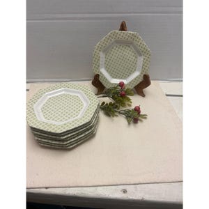 May include: A stack of six octagonal plates with a light green geometric pattern and white rims. One plate is displayed on a wooden stand, and a sprig of red berries adds a decorative touch. The plates are suitable for serving food.