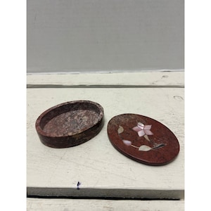 May include: Oval-shaped, open jewelry box with a matching lid. The box and lid are made of a reddish-brown stone. The lid features a flower design with pink and white accents. The box is open, revealing the interior.