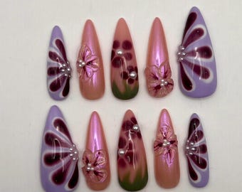 May include: A set of ten stiletto-shaped false nails. The nails feature a variety of designs, including iridescent pink, purple, and green hues. Some nails have 3D flower accents and pearl embellishments, while others showcase a floral pattern.