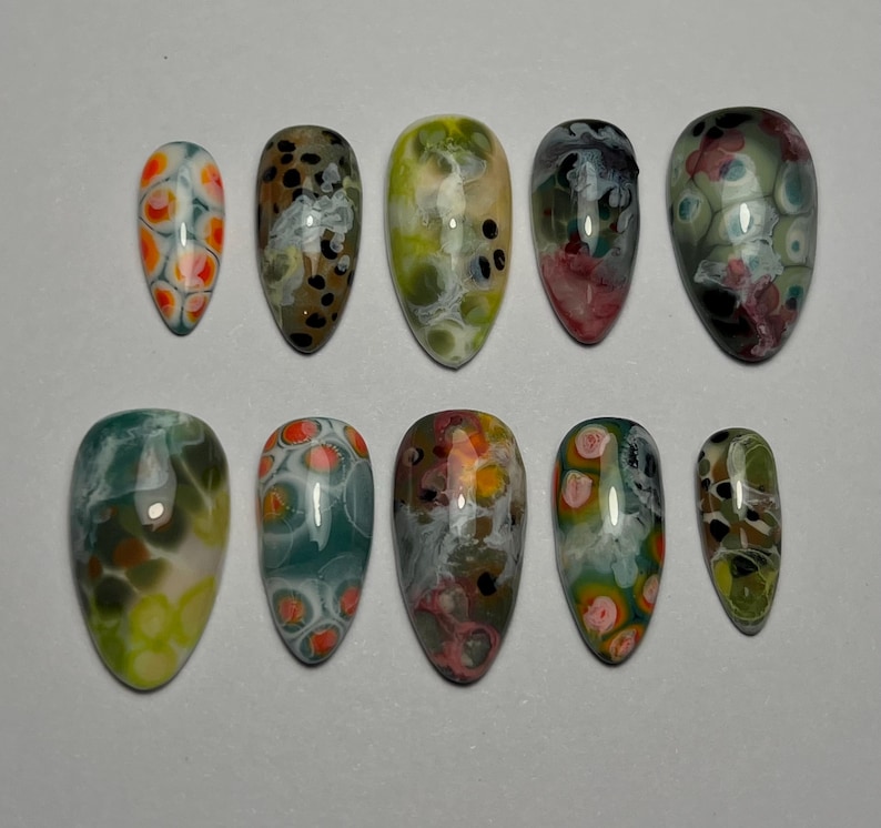 May include: A set of ten artificial nails with a glossy finish. Each nail features a unique abstract design with swirls of color, including orange, green, red, and black, against a white or clear base. The nails vary in size and shape.