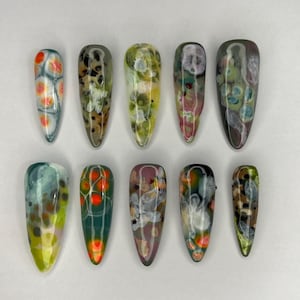 May include: A collection of ten long, almond-shaped artificial nails. Each nail features a unique abstract design with a glossy finish. Colors include orange, green, brown, white, and purple, creating a marbled effect. The nails are arranged on a white surface.
