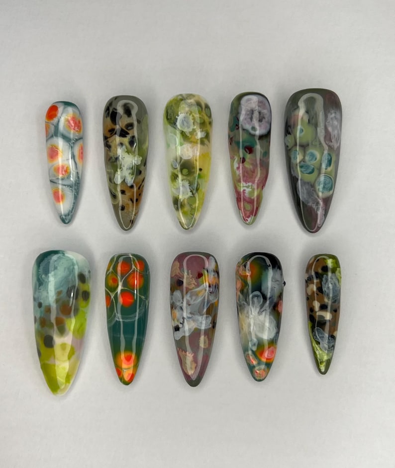 May include: A collection of ten long, almond-shaped artificial nails. Each nail features a unique abstract design with a glossy finish. Colors include orange, green, brown, white, and purple, creating a marbled effect. The nails are arranged on a white surface.