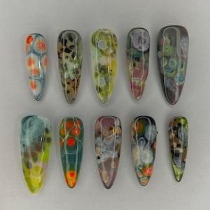 May include: Ten artificial nails with abstract designs. The nails are in various colors, including orange, green, yellow, and brown, with a glossy finish. The shapes are long and pointed, resembling a stiletto style.